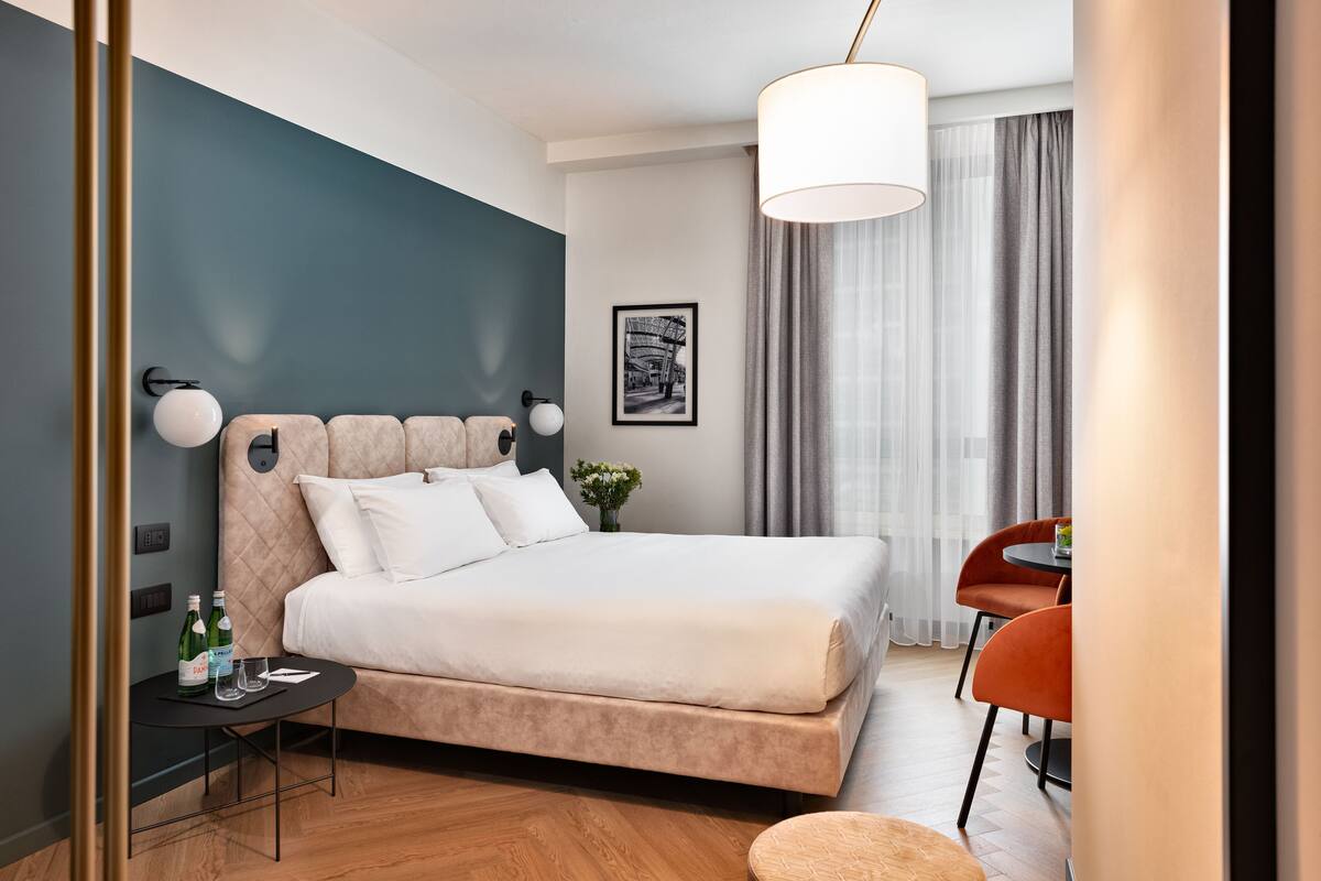 A modern hotel room with a large bed, a small table with a bottle and glass, a chair, and a dining area with a table and chairs.