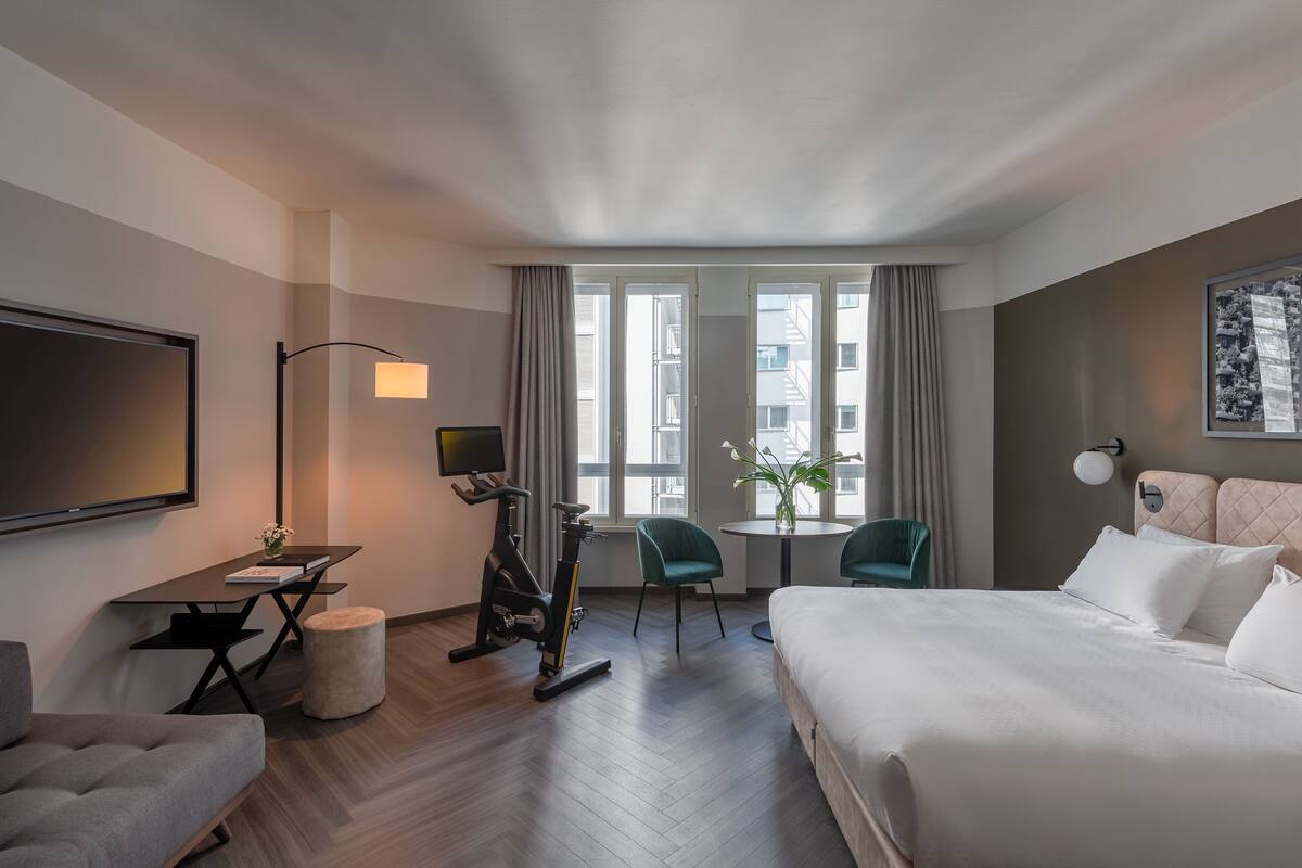 A hotel room with a bed, a desk, an exercise bike, two chairs, a TV, and a window with curtains.