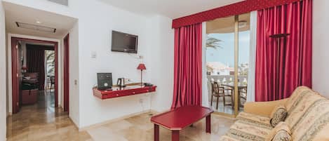 Habitación de hotel con sofá, una mesita, televisor montado en la pared y puerta corrediza de vidrio que da a un balcón.