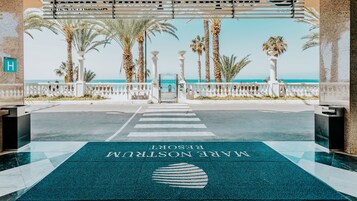 Entrada de hotel com vista para a praia e palmeiras.