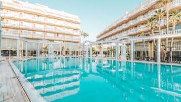 Una gran piscina al aire libre rodeada de edificios hoteleros de varios pisos con balcones y palmeras.