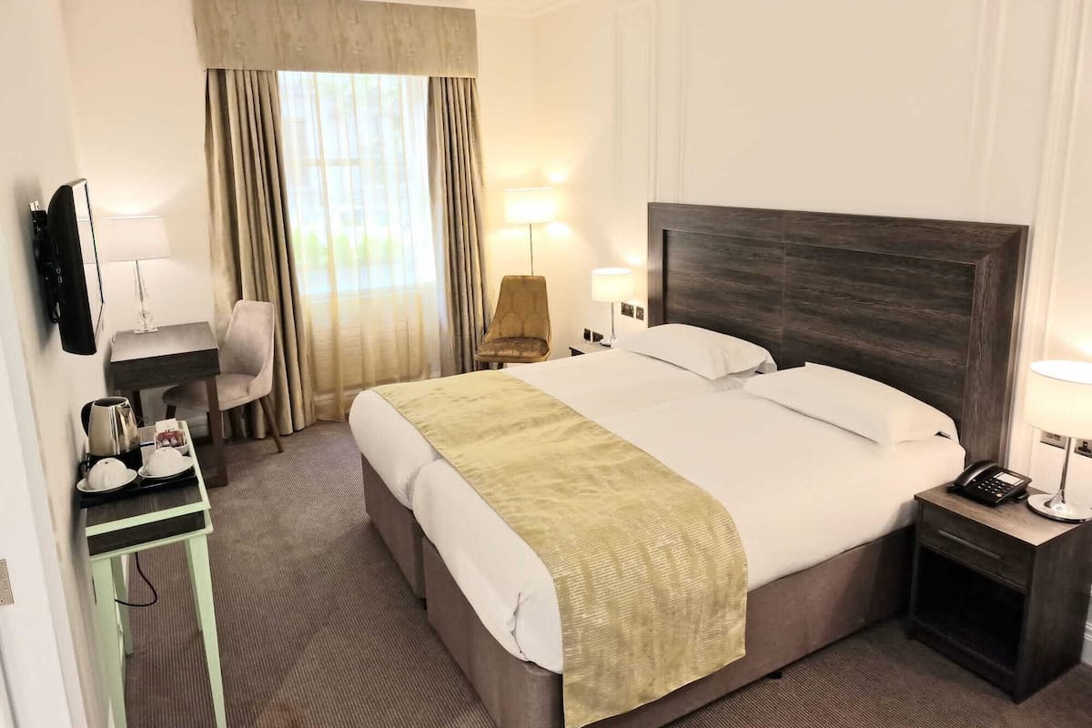 A hotel room with a large bed, a desk with a chair, a TV, and a bedside table with a lamp.