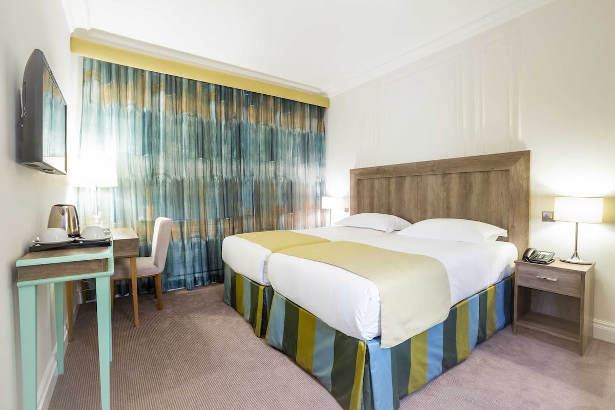 A hotel room with a bed, a desk with a chair, a nightstand with a lamp, and a large window with curtains.