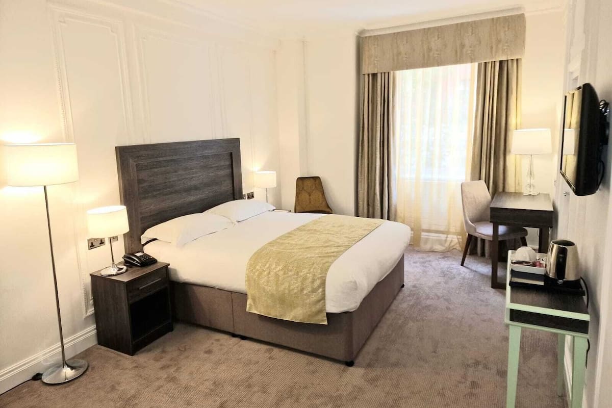 A hotel room with a large bed, bedside lamps, a desk with a chair, a TV, and a mirror.