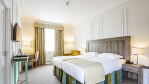 A hotel room with a large bed, a desk with a chair, a TV, and a bedside table with a lamp. - Astor Court Hotel (London)