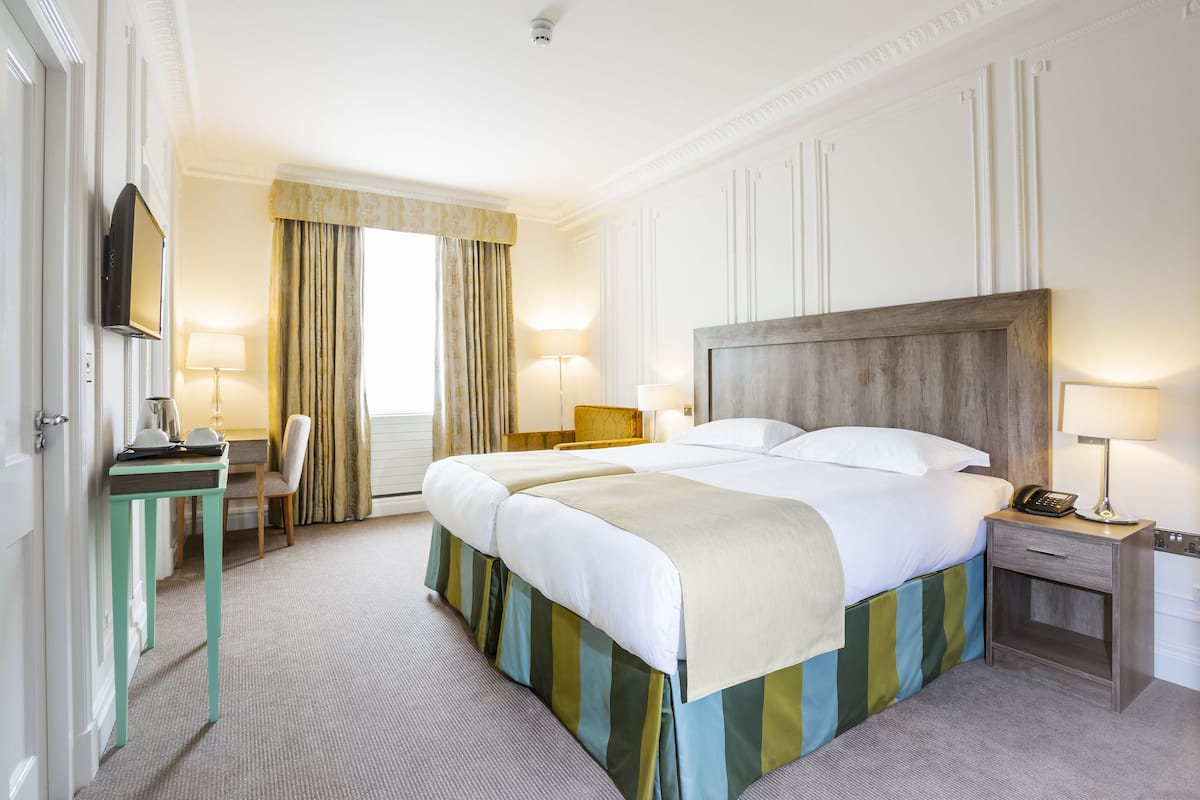 A hotel room with a large bed, a desk with a chair, a TV, and a bedside table with a lamp.