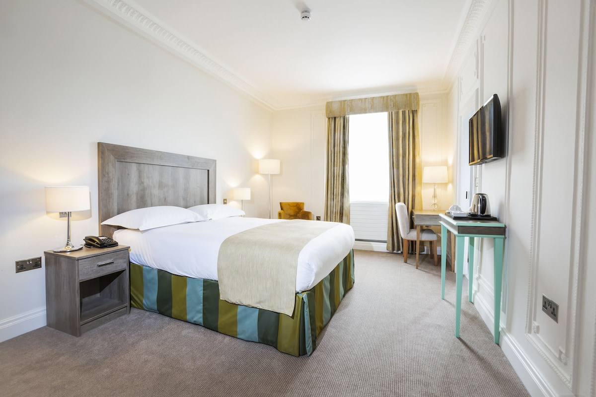 A hotel room with a bed, bedside table, lamp, desk, chair, and TV.