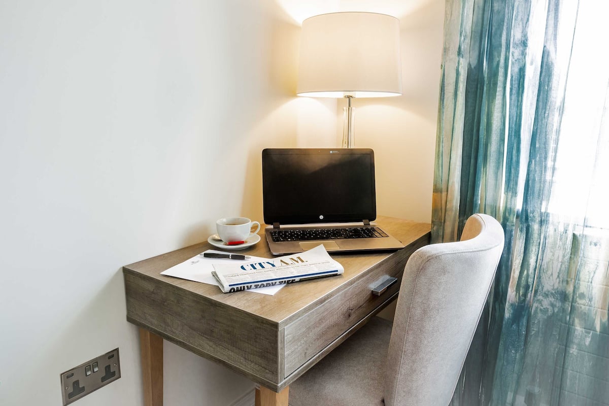 A hotel room with a wooden desk, a laptop, a cup of coffee, and a magazine.