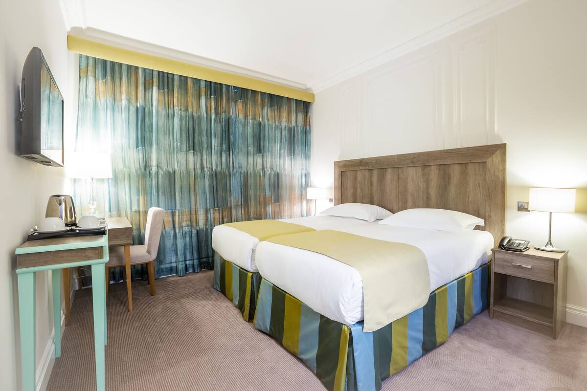 A hotel room with a bed, a desk with a chair, a TV, a bedside table with a lamp, and a curtain with a yellow trim.