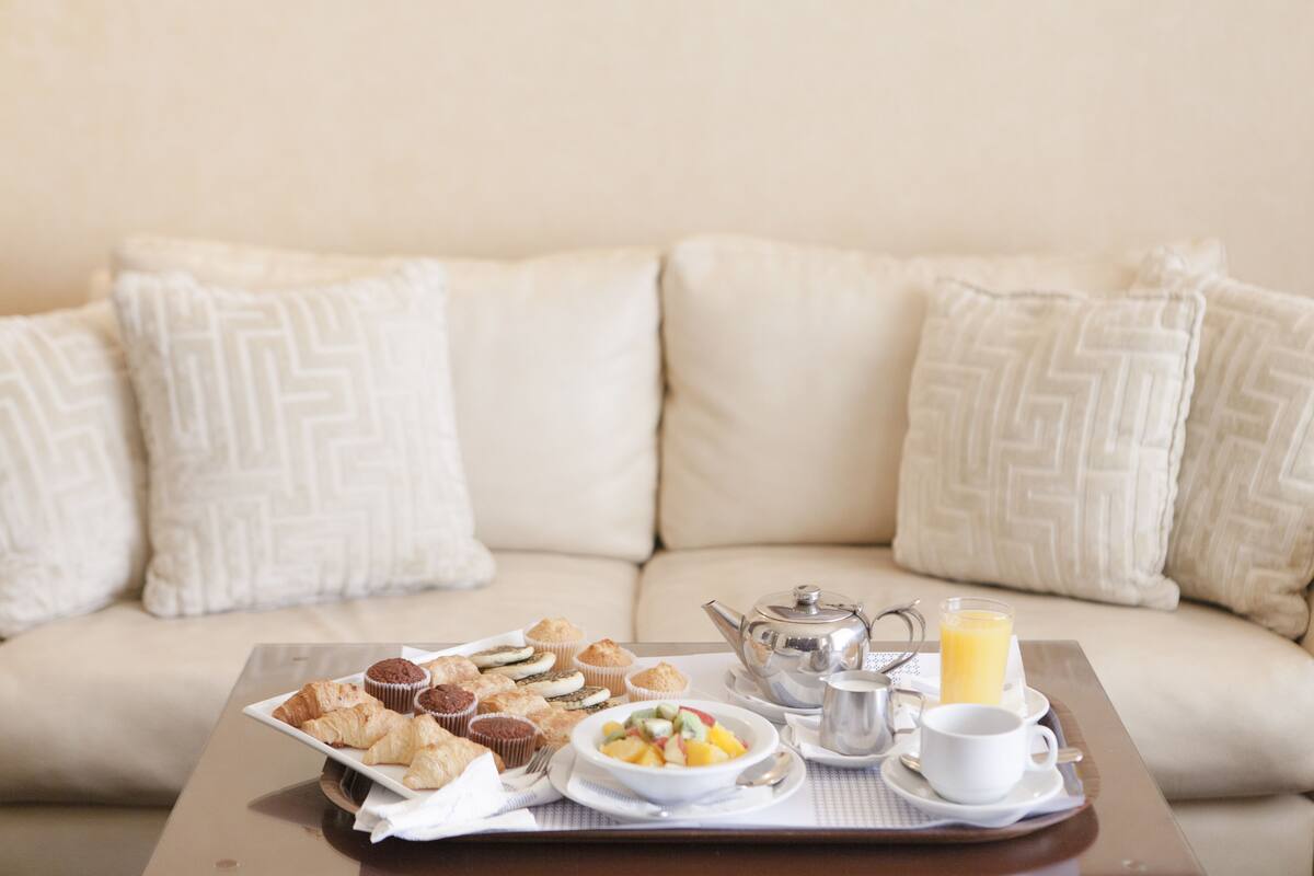 A breakfast tray with pastries, a bowl of fruit, a teapot, a cup, and a glass of orange juice on a tray, placed on a table in front of a beige sofa with patterned cushions.
