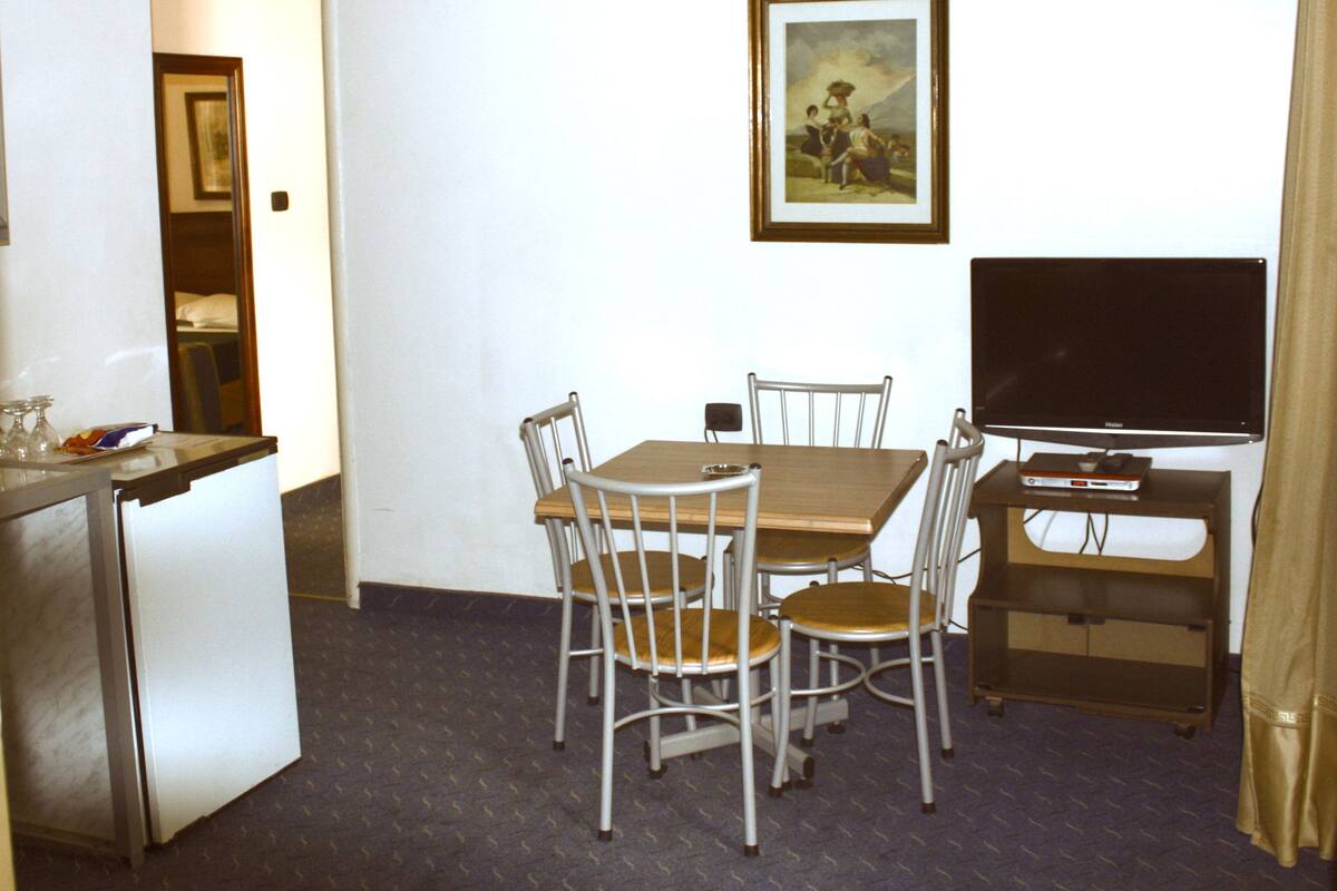 A compact hotel room with a dining table, four chairs, a television, and a small refrigerator.
