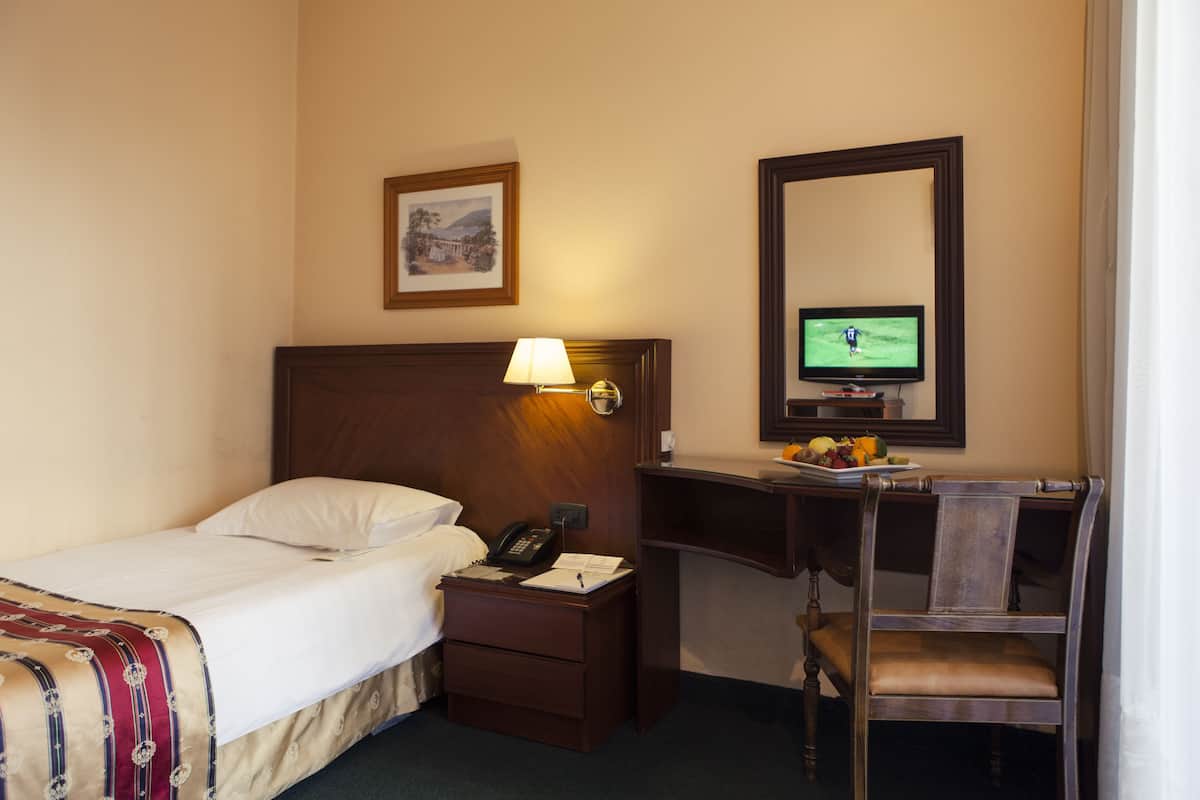 A hotel room with a bed, bedside table with a lamp, a desk with a computer monitor, and a chair.