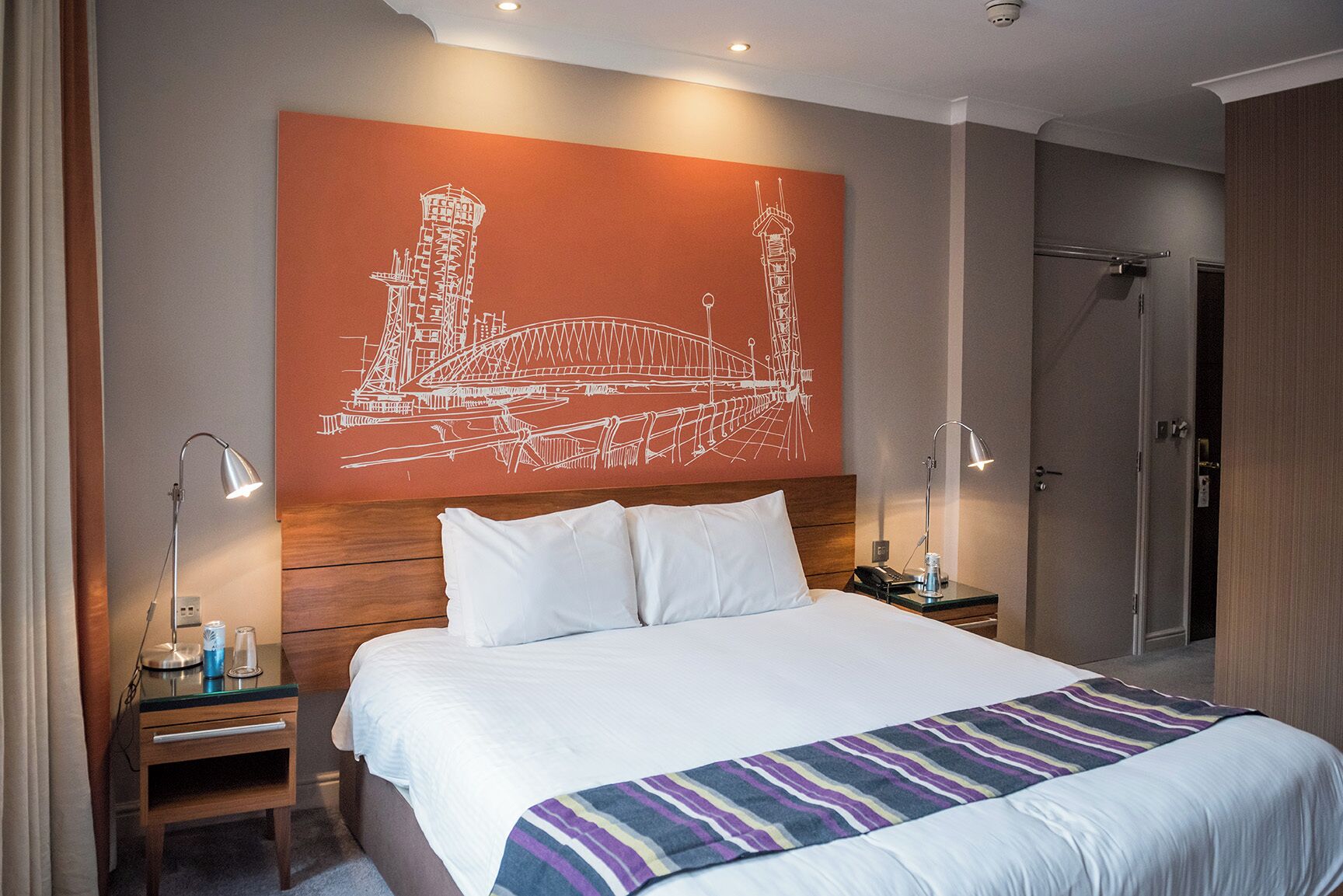 A hotel room with a large bed, a nightstand with a lamp, a bedside table with a phone, and a wall art of a bridge.