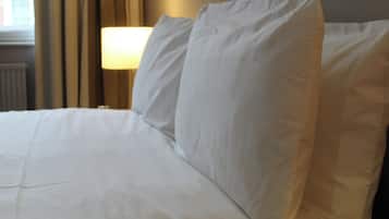 Una cama bien hecha con sábanas y almohadas blancas, una lámpara al lado y vista a un edificio a través de una ventana con cortinas.