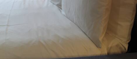 Una cama bien hecha con sábanas y almohadas blancas, una lámpara al lado y vista a un edificio a través de una ventana con cortinas.