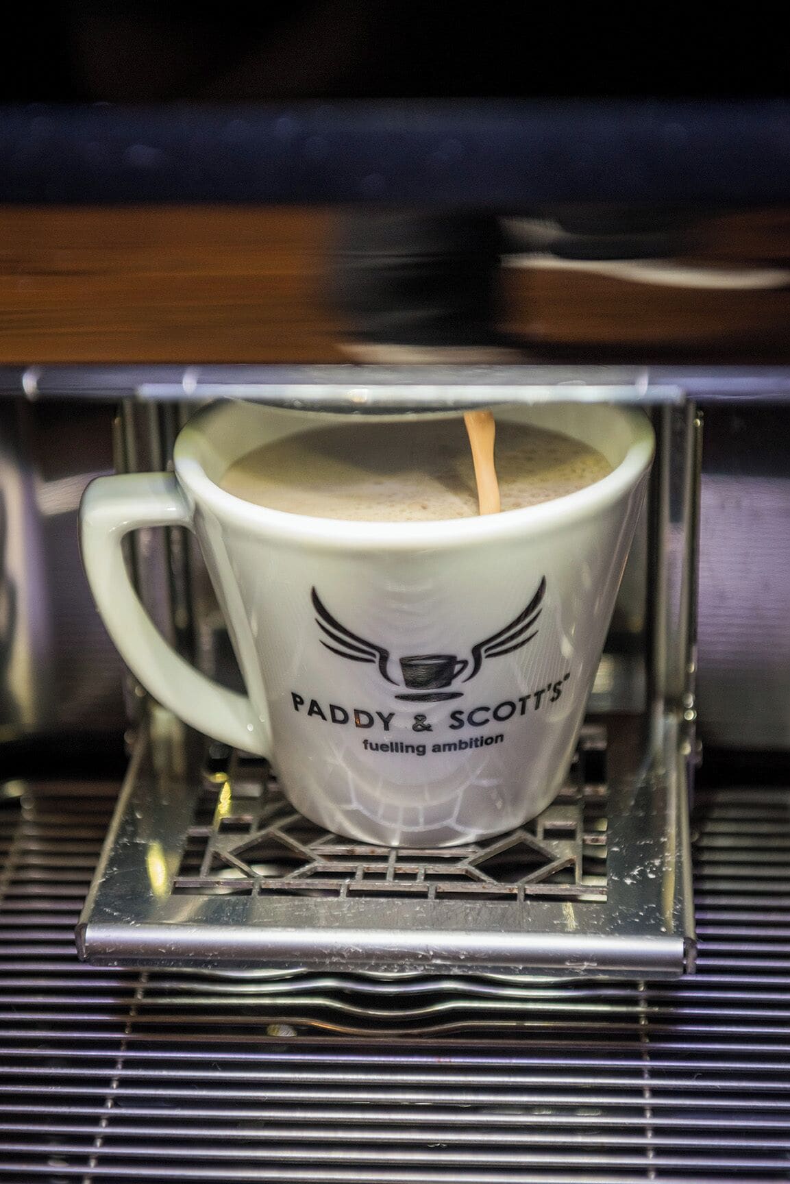 Eine weiße Kaffeetasse mit dem Aufdruck "Paddy & Scott's fuelling ambition" steht auf einem Metallgestell in einer Maschine.