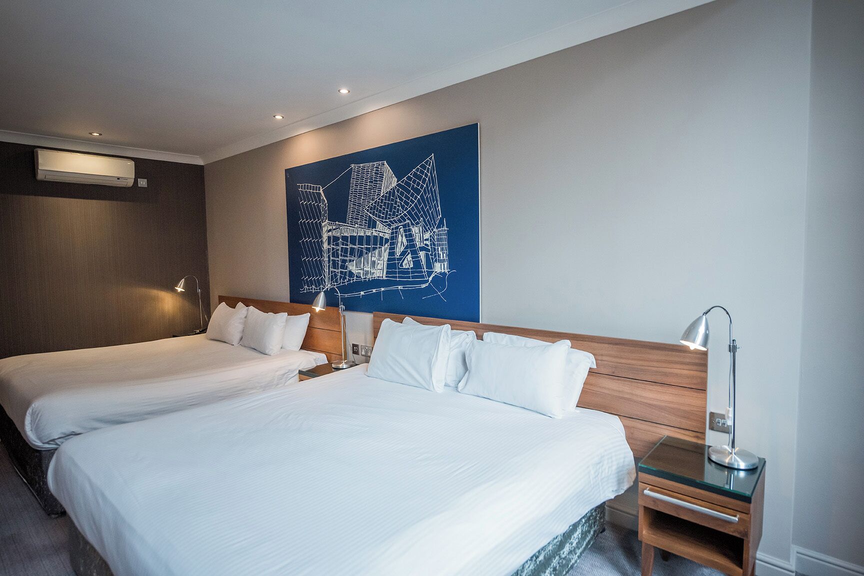 Ein Hotelzimmer mit zwei Betten, einem hölzernen Kopfteil, einem Nachttisch mit Lampe und einem großen, abstrakten Gemälde an der Wand.