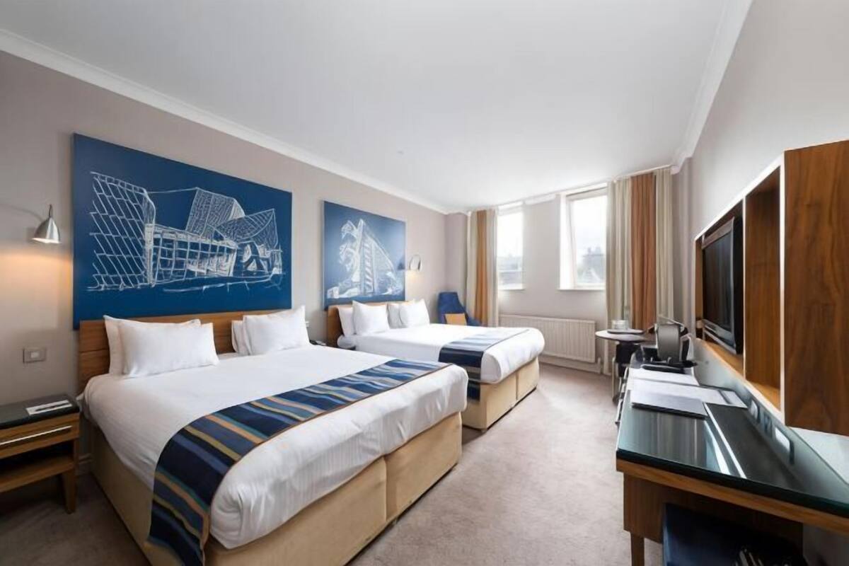 A hotel room with two beds, a desk, a TV, and large windows.