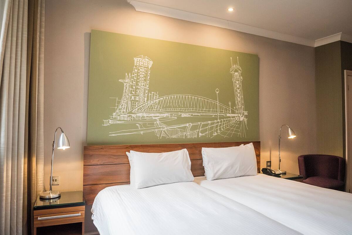 A hotel room with a large bed, two bedside tables, a nightstand, a lamp, and a wall mural of a bridge.