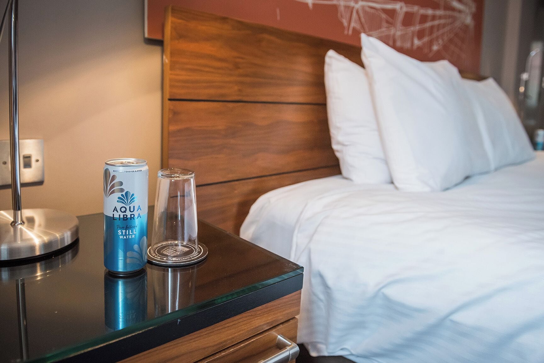 Quarto de hotel com cabeceira de madeira, cama com roupa de cama branca, mesa de vidro com uma lata de água Aquafina e um copo.