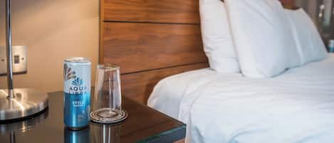 Quarto de hotel com cabeceira de madeira, cama com roupa de cama branca, mesa de vidro com uma lata de água Aquafina e um copo.