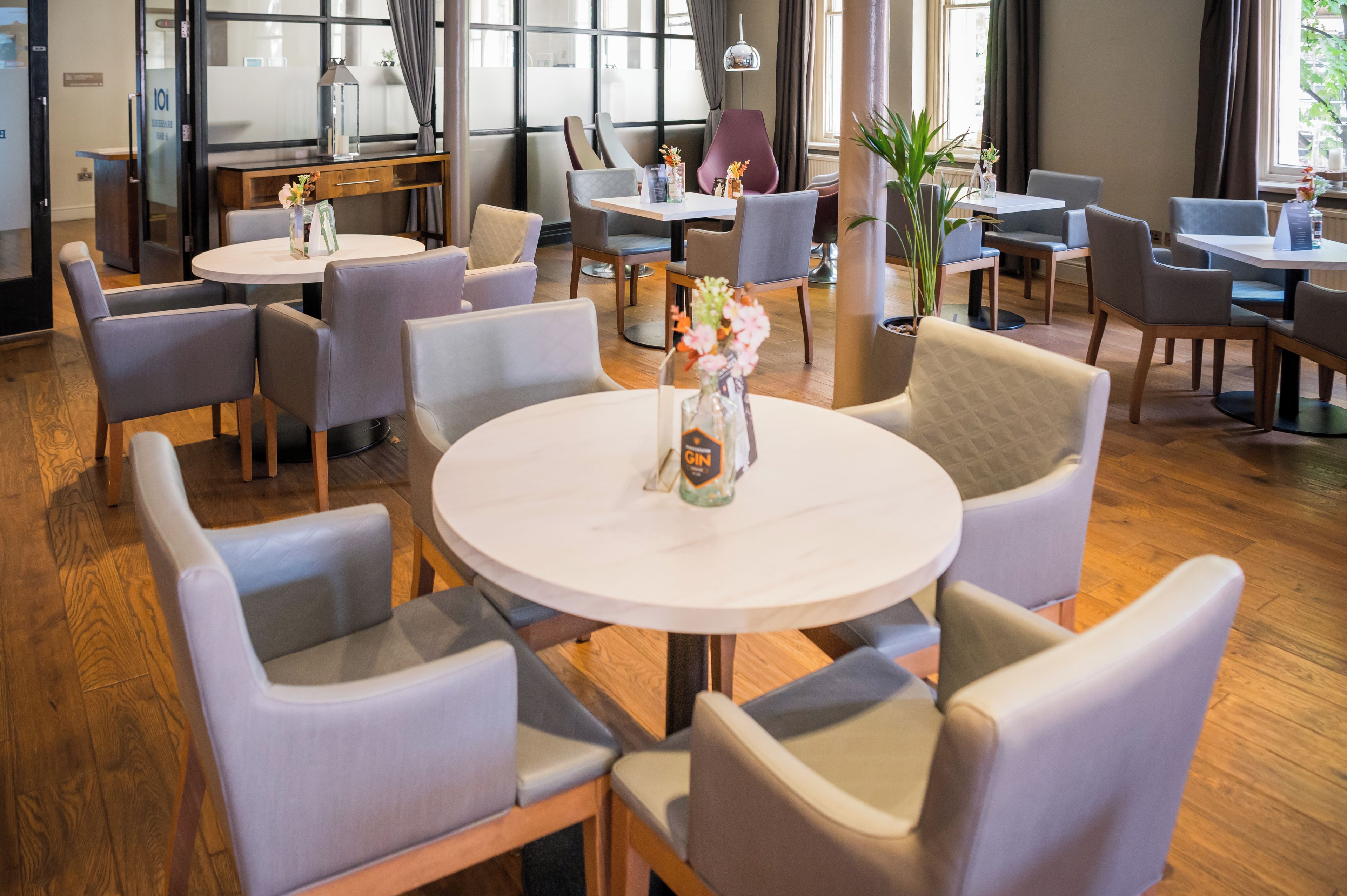 A hotel dining area with round tables and cushioned chairs, a potted plant, and a bottle of gin on the tables.