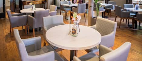 A hotel dining area with round tables and cushioned chairs, a potted plant, and a bottle of gin on the tables.