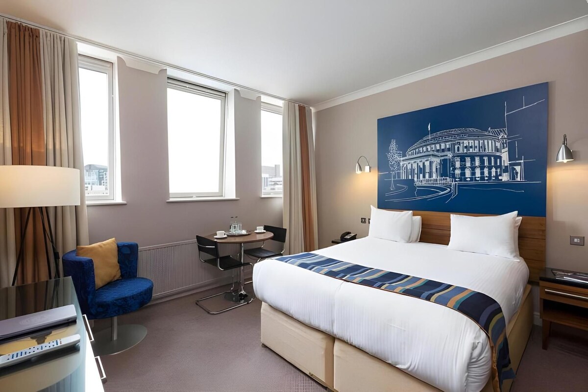 A hotel room with a large bed, a desk, a chair, a lamp, and a painting on the wall.
