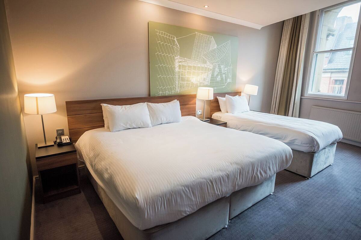 A hotel room with two beds, a nightstand with a lamp, a telephone, and a wall art piece.