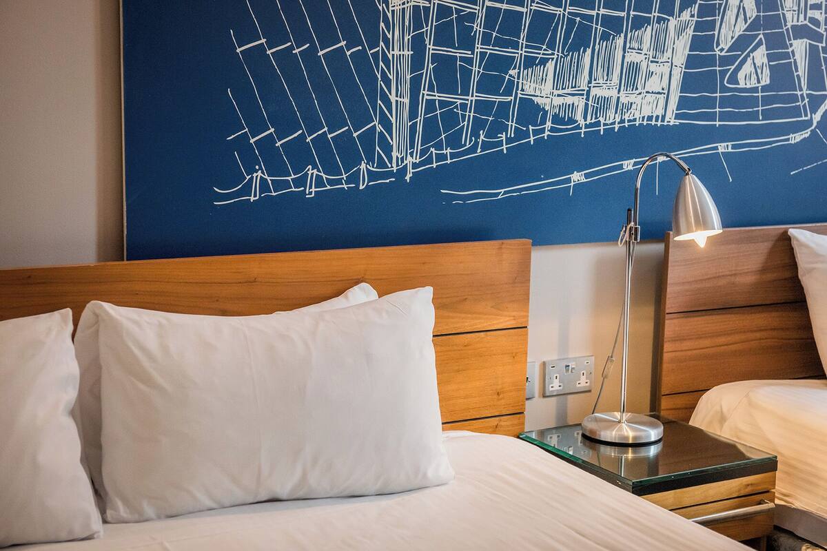 A hotel room with a wooden headboard, white bedding, a glass bedside table with a lamp, and a wall with a geometric design.