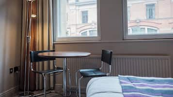 A hotel room with a bed, a small round table, two chairs, a floor lamp, and a window with a view of buildings.