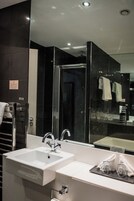 Un baño moderno con un lavabo blanco, cabina de ducha con mampara de vidrio y un espejo que refleja un perchero y un lavabo.