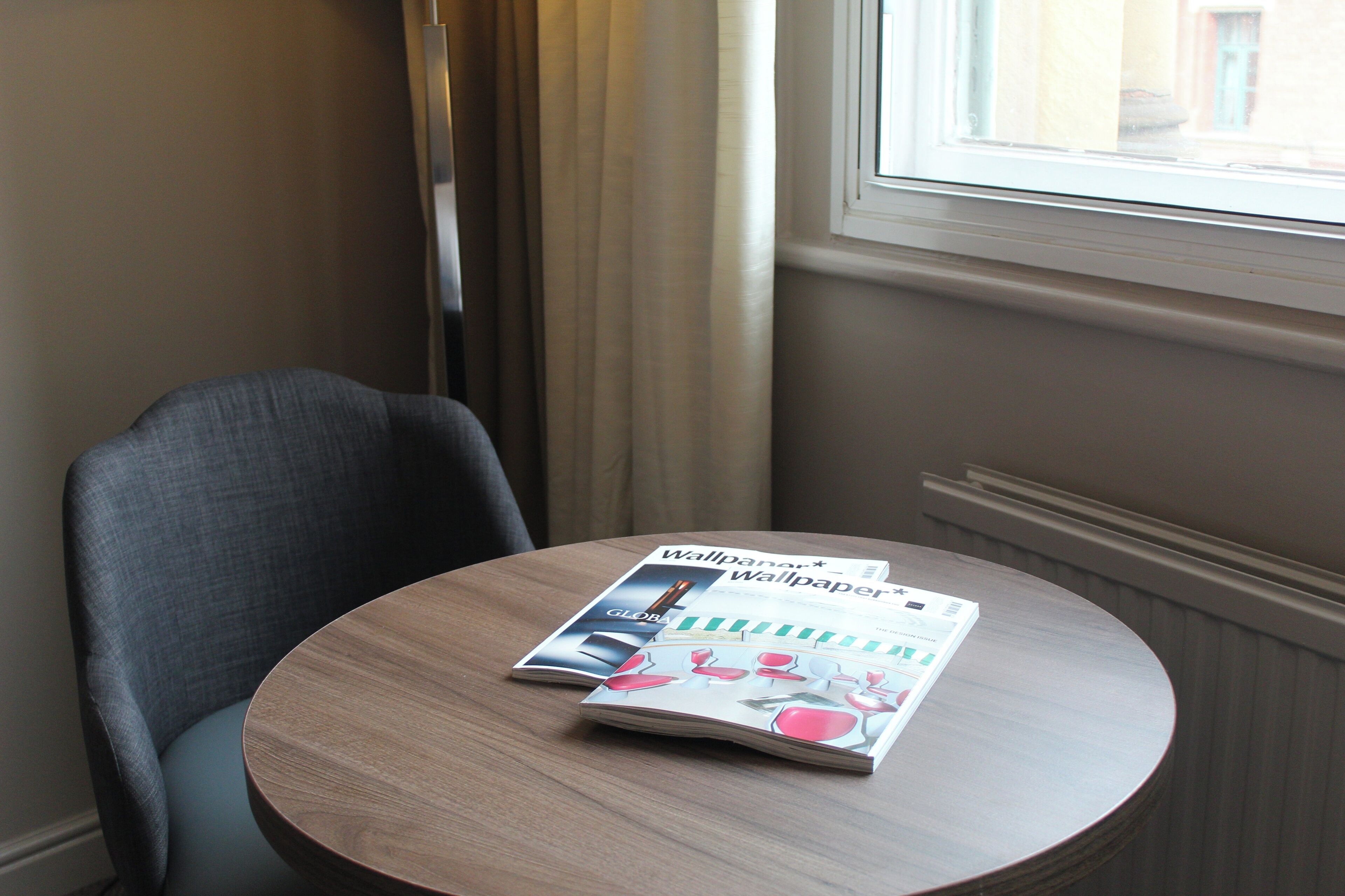 A round wooden table with a magazine on it, a grey upholstered chair, and a window with curtains.