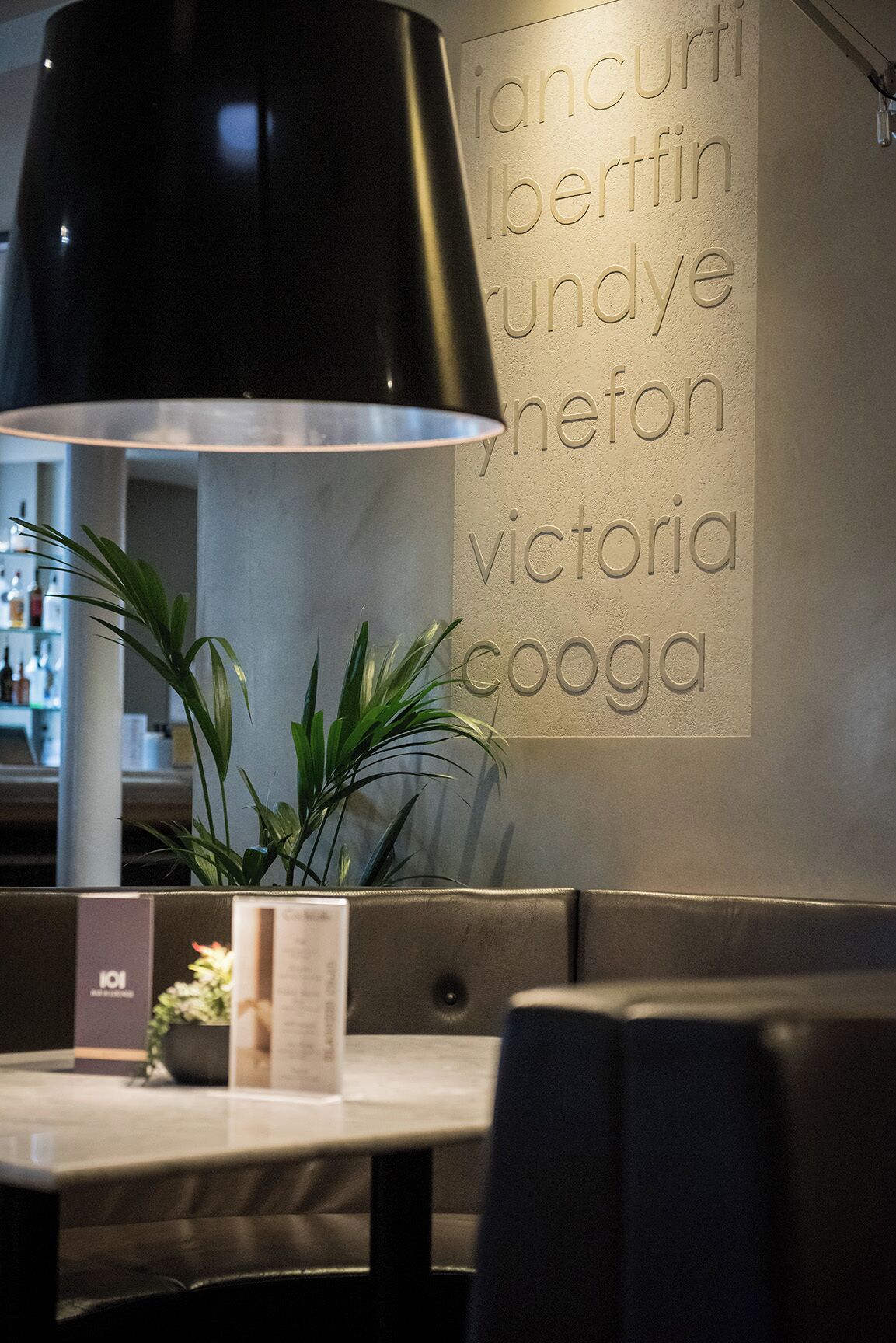 Interior de restaurant amb taula, cadires, una làmpada negra i una paret amb text relleu.