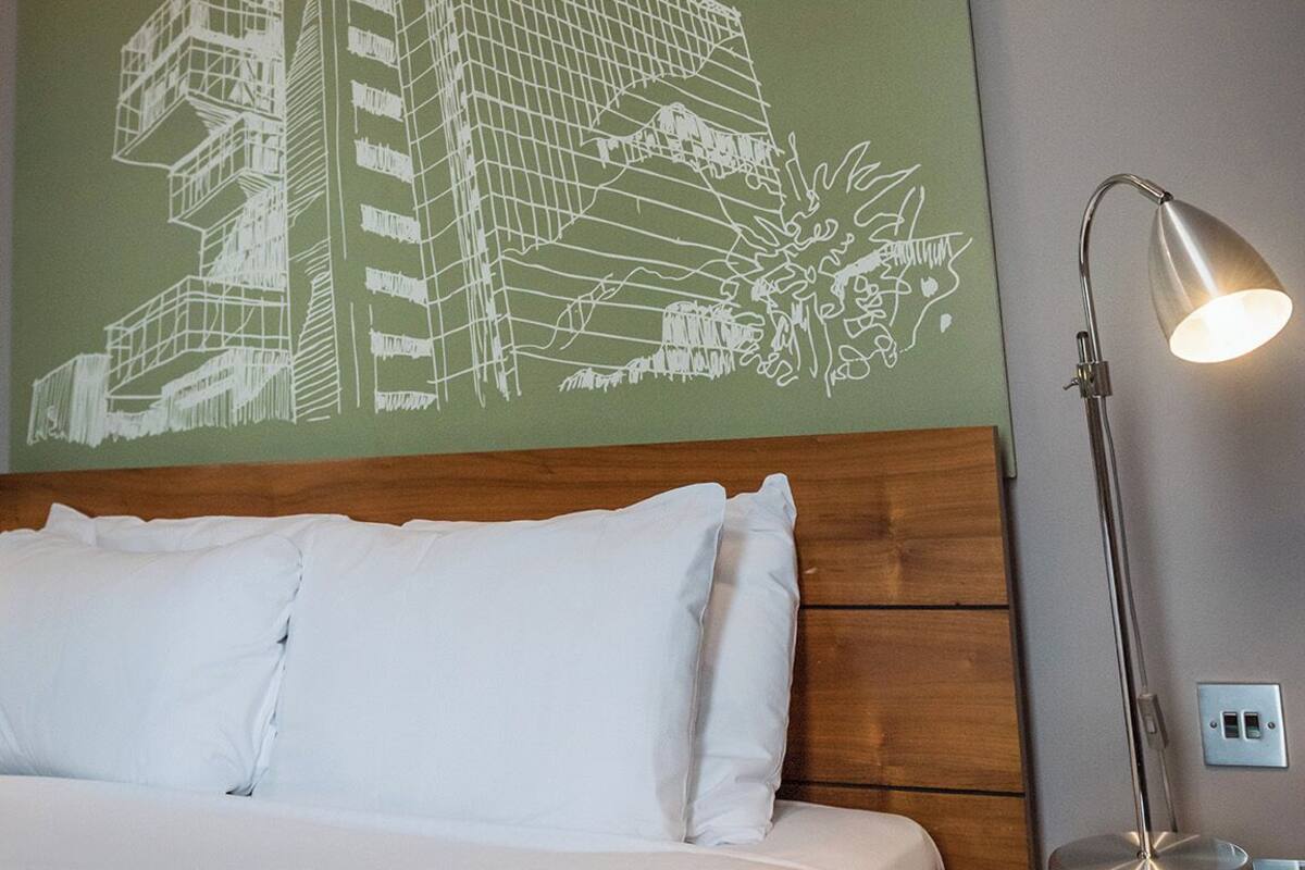 A hotel room with a bed, a nightstand, a lamp, and a wall with a building outline.