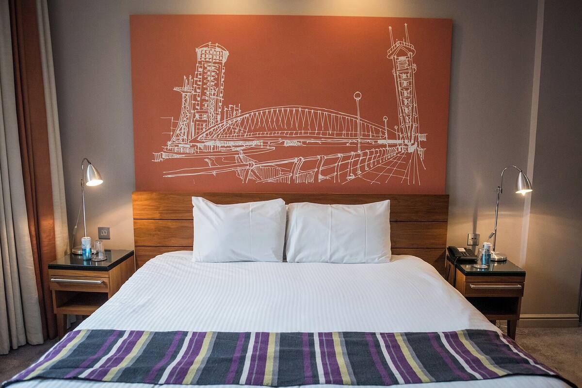 A hotel room with a large bed, two bedside tables, a nightstand, a striped area rug, and a wall art of a bridge.
