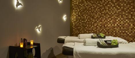 Ein Spa-Raum mit zwei Massagetischen, einer Holzwand mit Pixelmuster und einem Regal mit Flaschen und Kerzen.