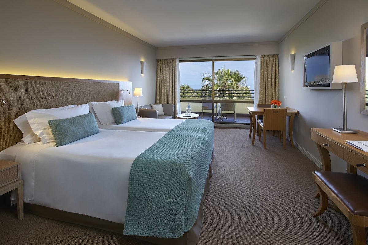 A hotel room with two beds, a desk, a chair, a TV, and a balcony with a view.