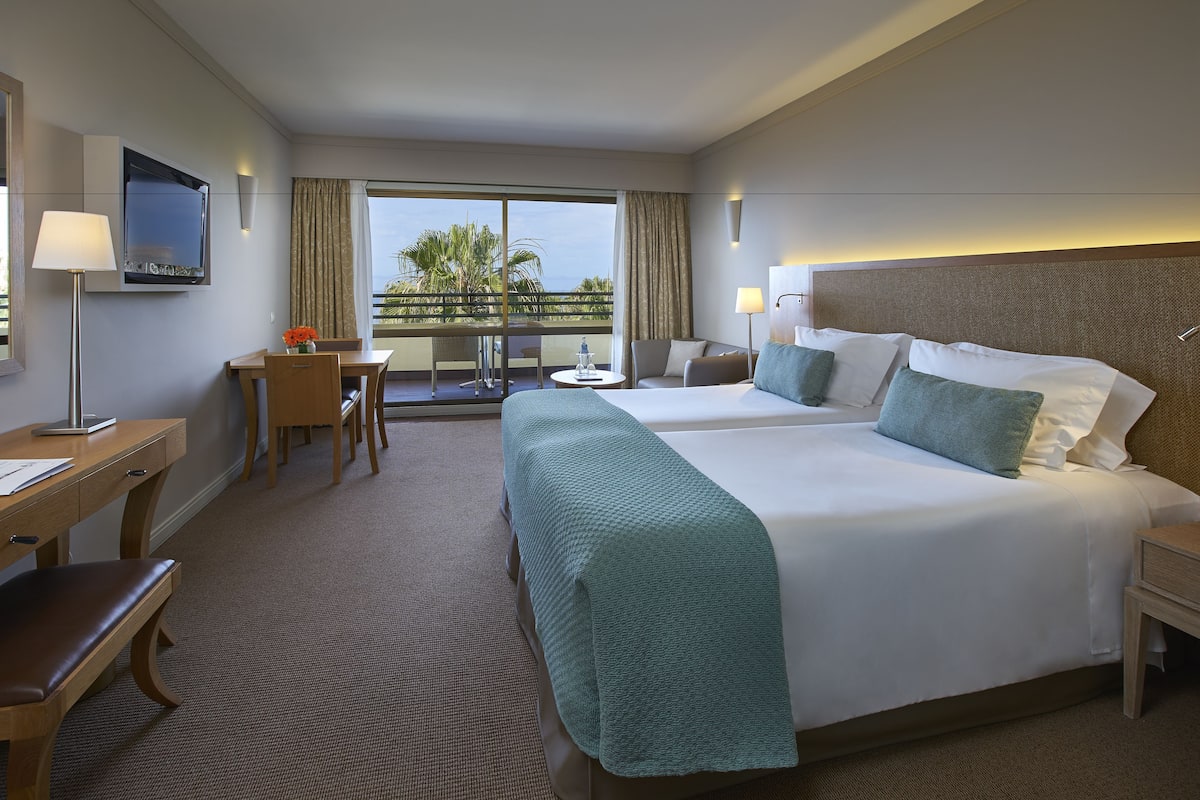 A hotel room with a large bed, a desk, a chair, a small table, and a balcony with a view of palm trees.