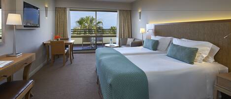 A hotel room with a large bed, a desk, a chair, a small table, and a balcony with a view of palm trees.