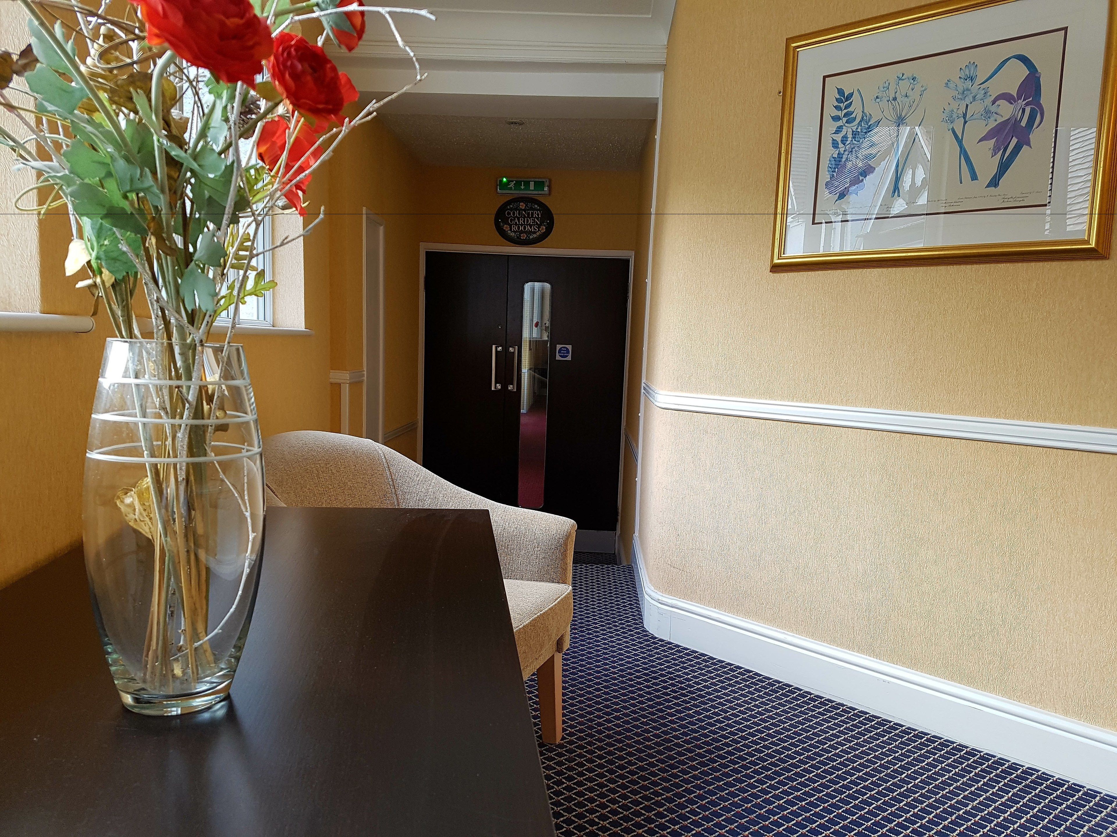 Une chambre d’hôtel avec une table en bois foncé, un vase rempli de fleurs, une illustration botanique encadrée et une porte portant l’inscription « Chambres Country Garden ».