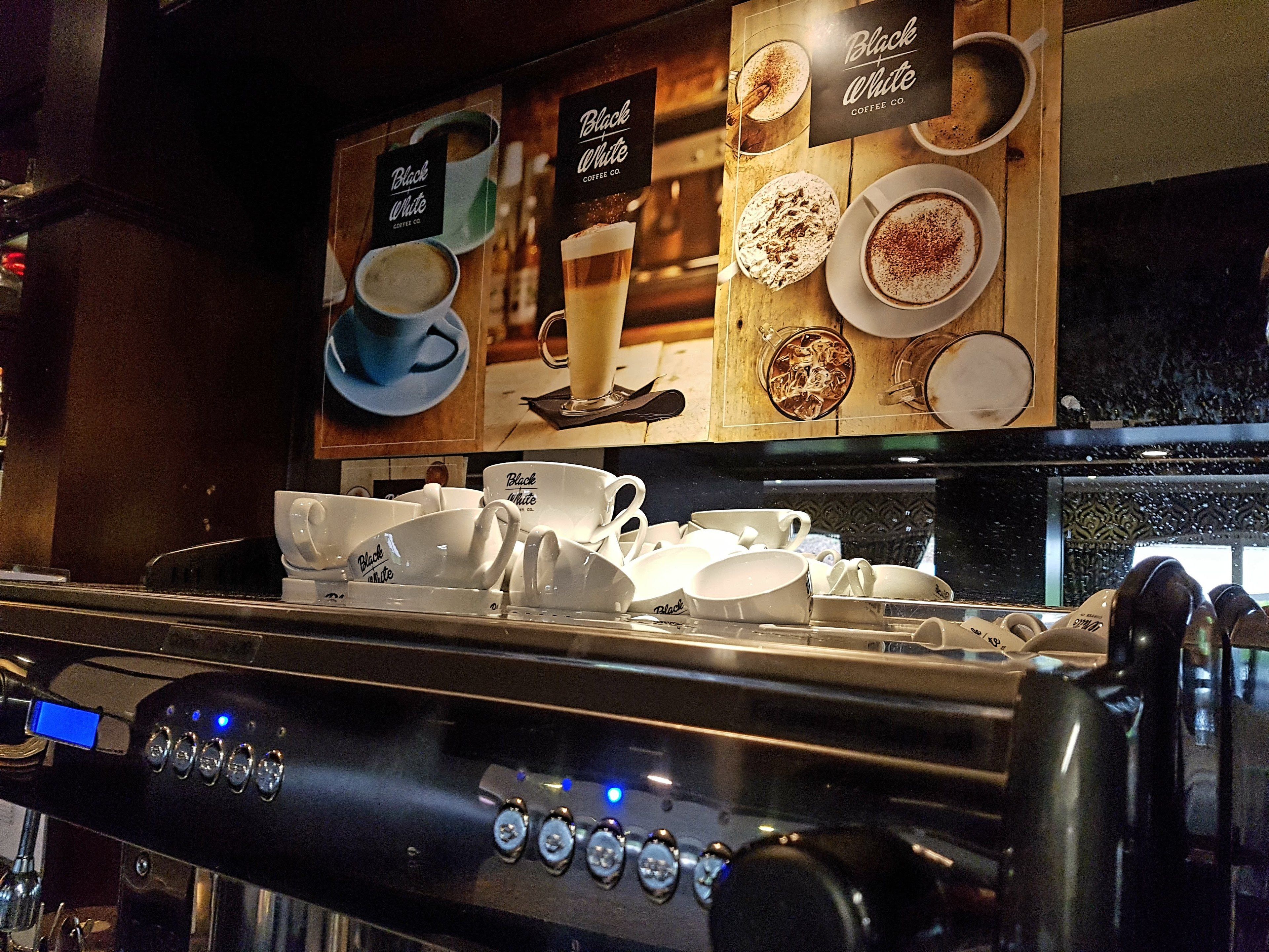A coffee machine with a digital display and control buttons, a coffee menu above it, and a stack of white cups and saucers below.