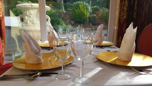 Mesa lista para una comida, con copas transparentes y platos dorados, ubicada cerca de una ventana con vistas a follaje y una fuente.