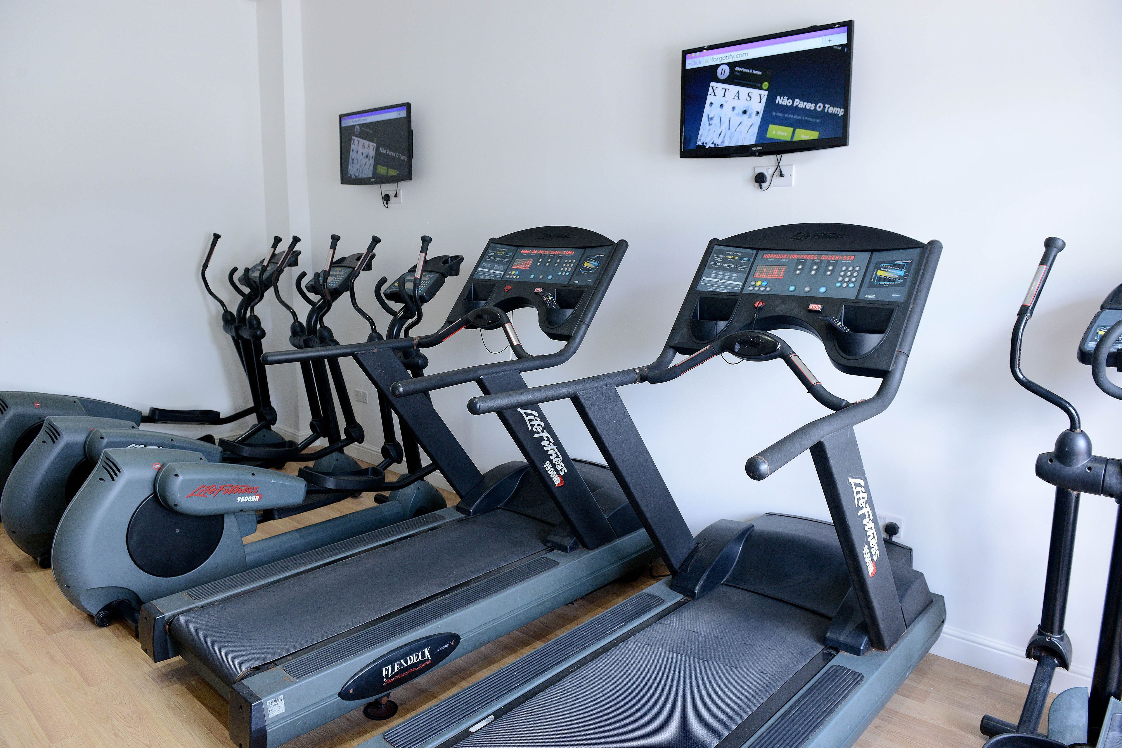Une salle de sport équipée de plusieurs tapis de course et vélos d’exercice, d’un écran numérique et d’un écran fixé au mur.