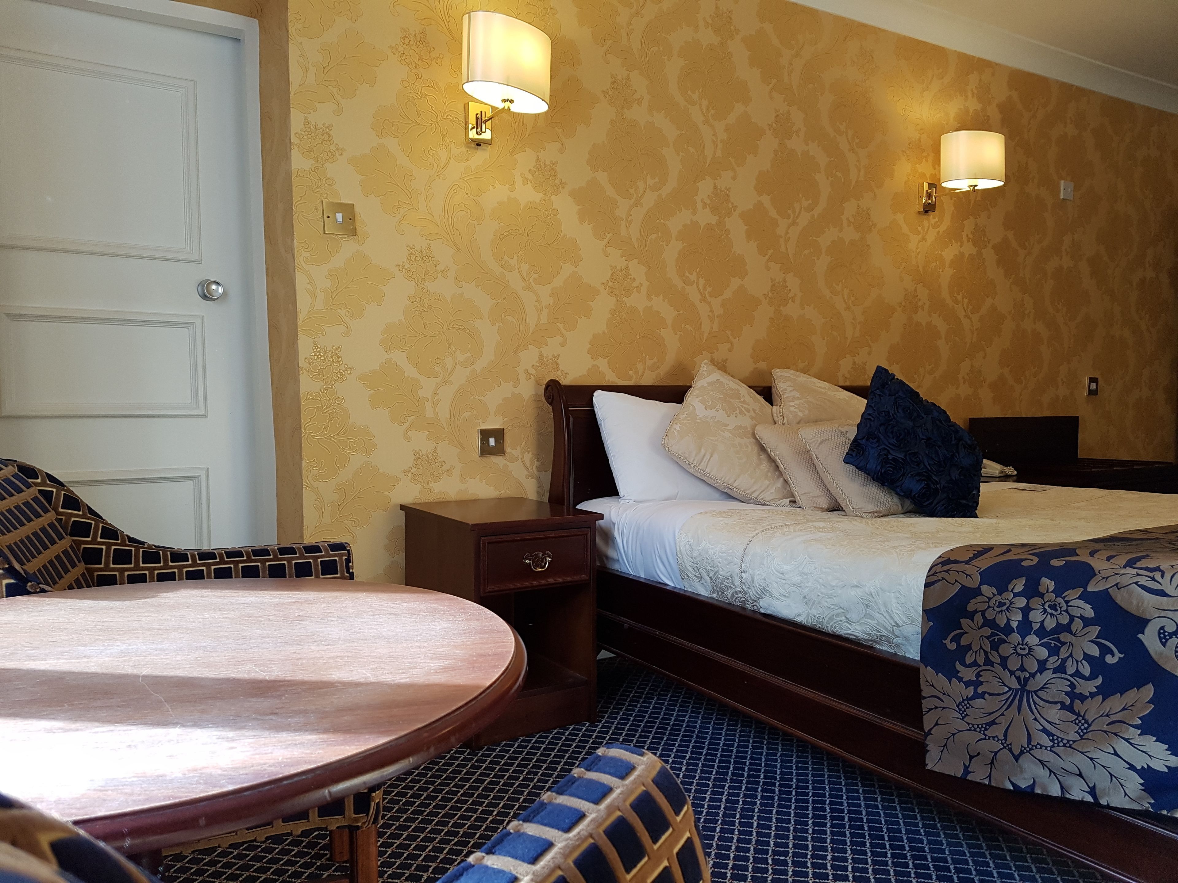 A hotel room with a bed, bedside table, lamp, and a round wooden table.