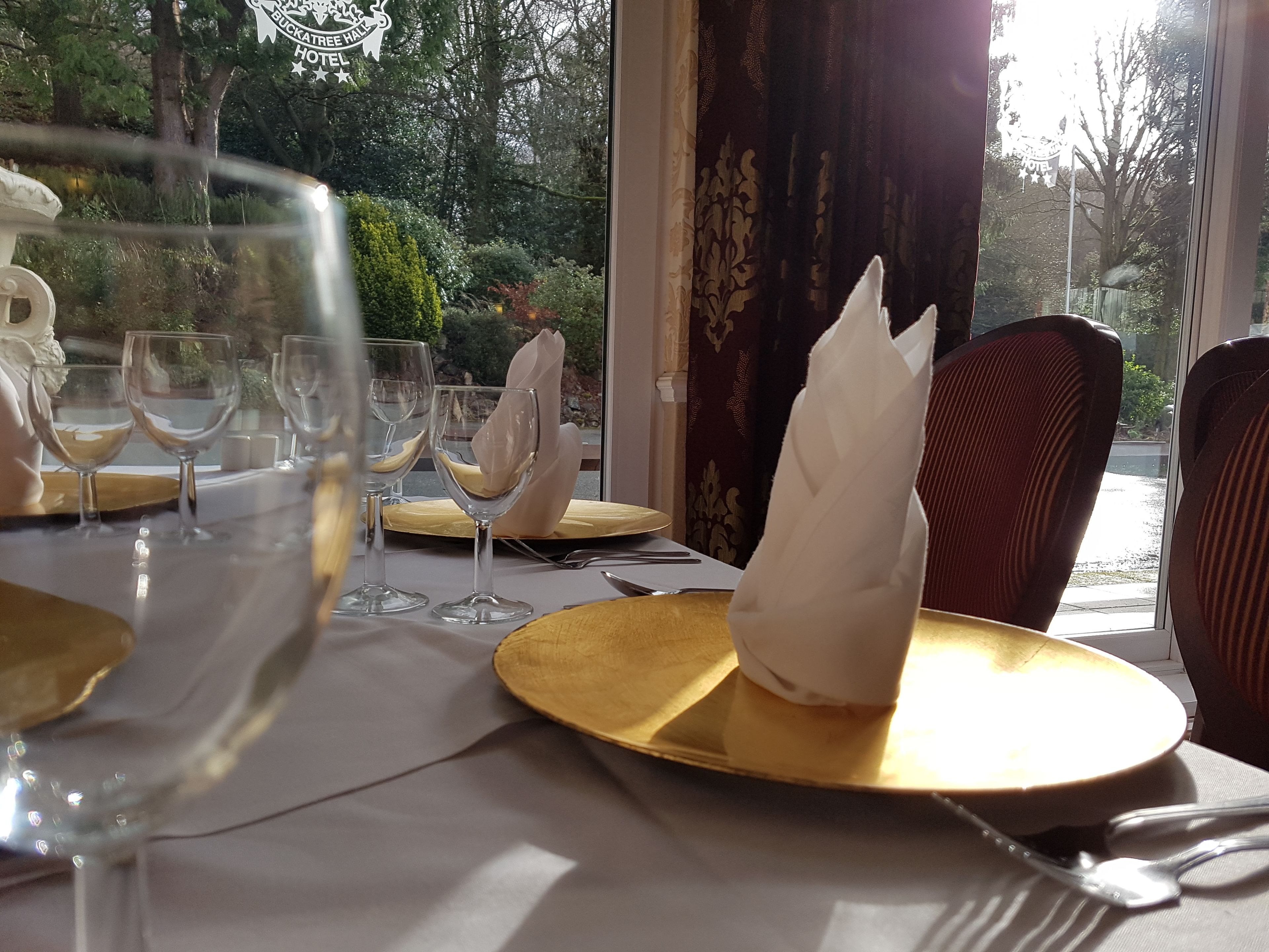 A dining table set with wine glasses, plates, and cutlery, with a view of trees and a sign reading "Buckatree Hall Hotel".