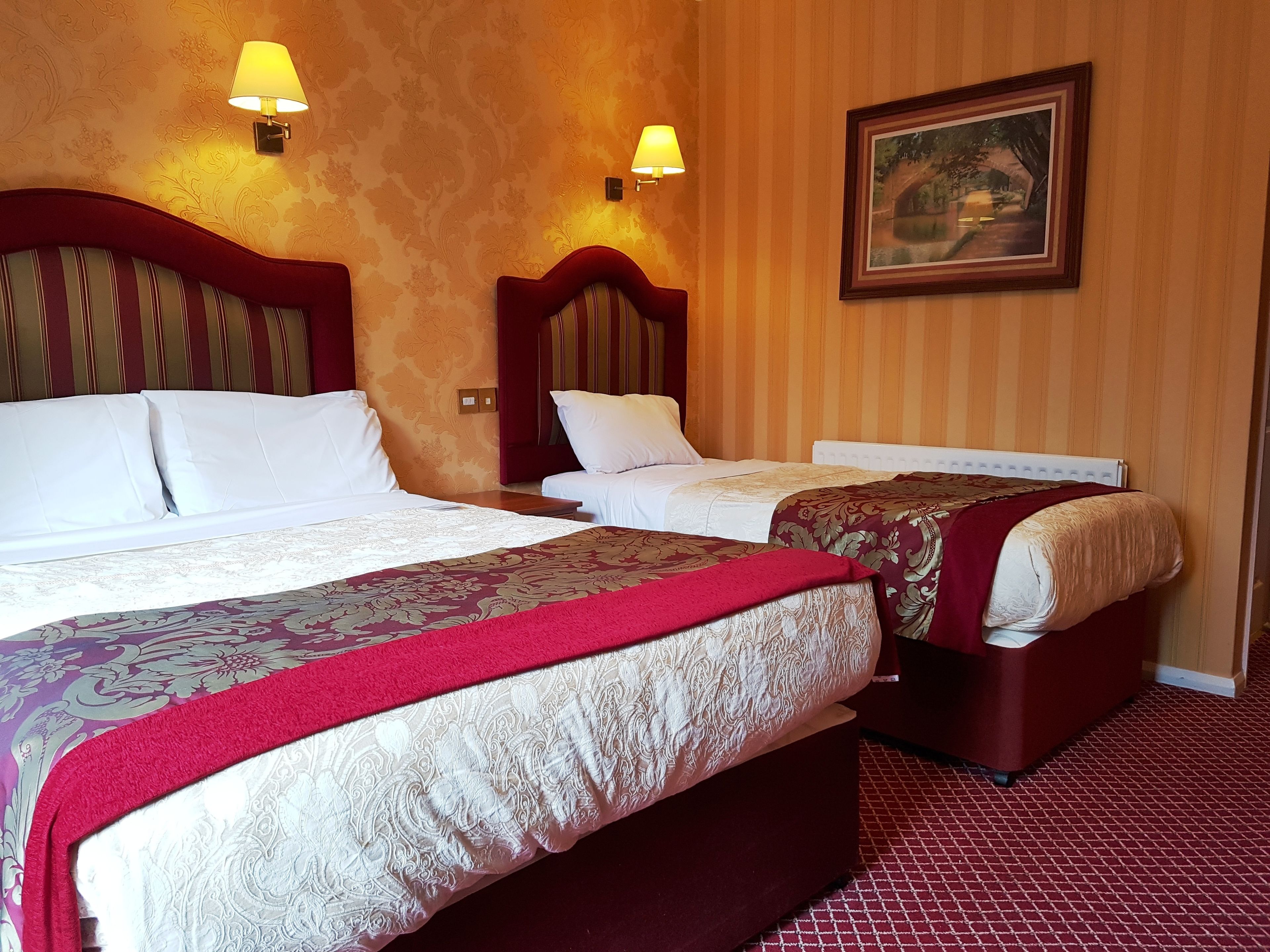 A hotel room with two beds, each with a headboard, white bedding, and a red throw blanket. There are wall-mounted lamps and a framed picture on the wall.