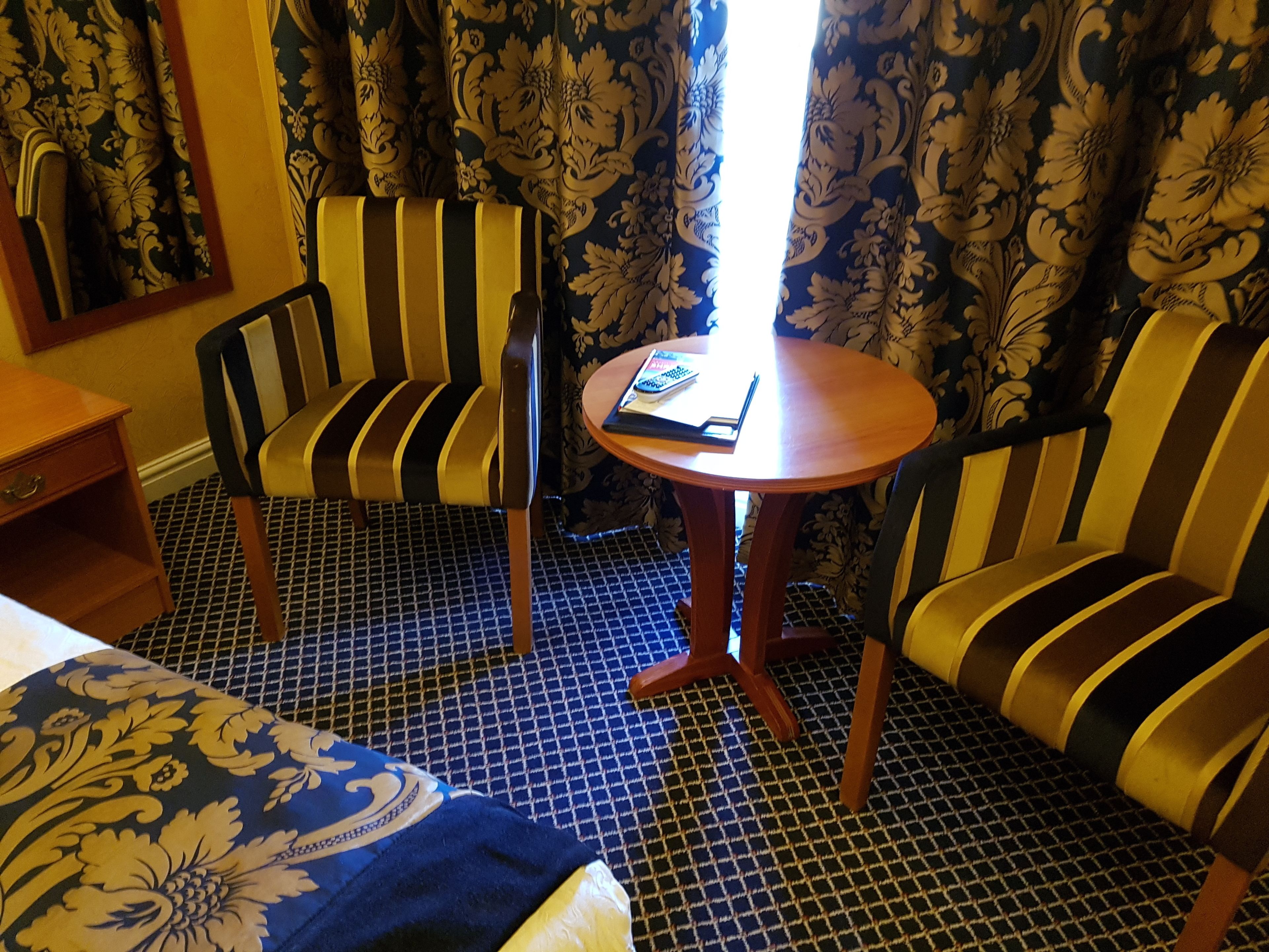 A hotel room with a bed, two striped armchairs, a small wooden table, and a patterned carpet.