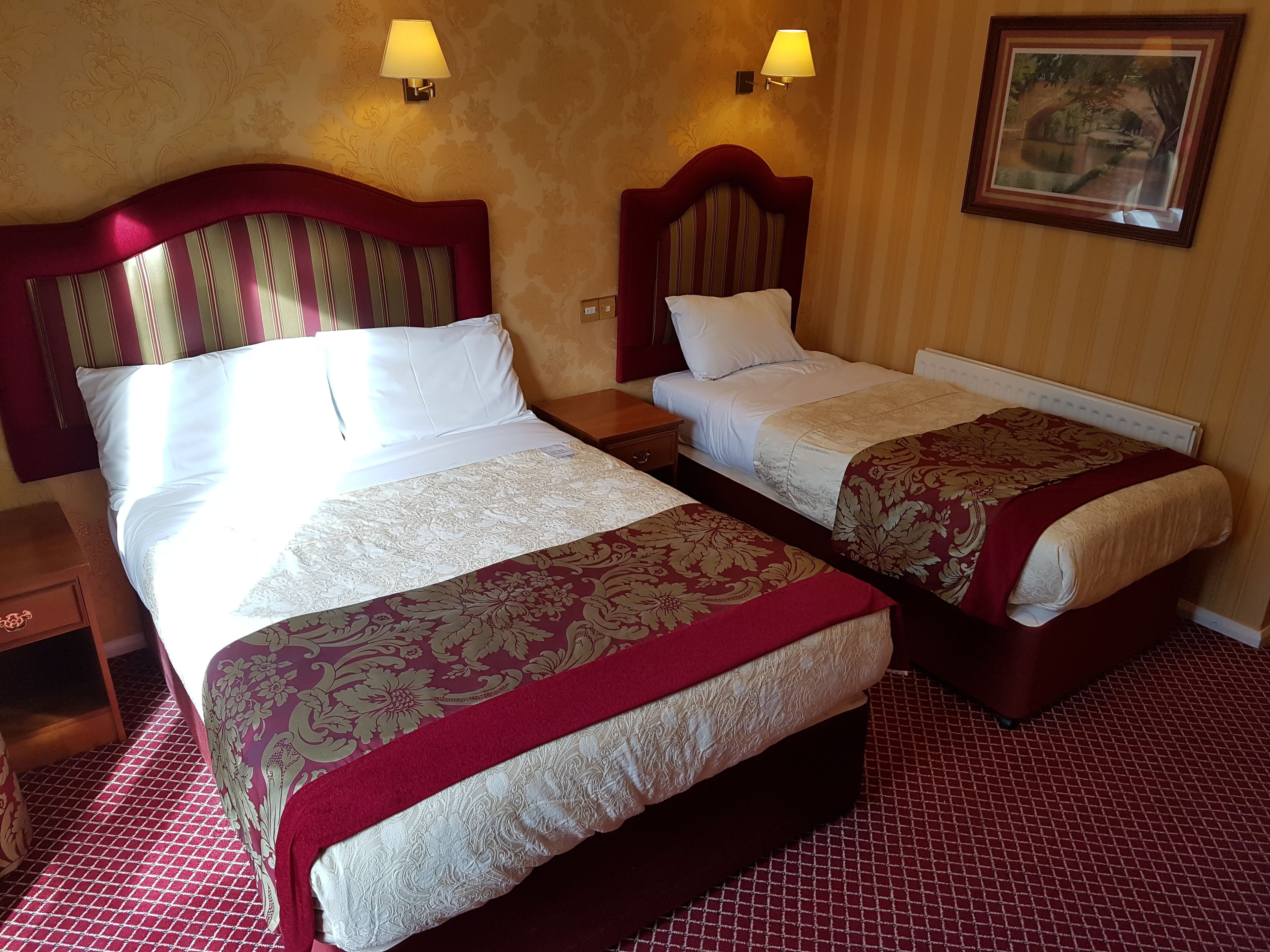 A hotel room with two beds, each with a wooden headboard and a red and white bedspread. There are two bedside tables with lamps, and a framed picture on the wall.