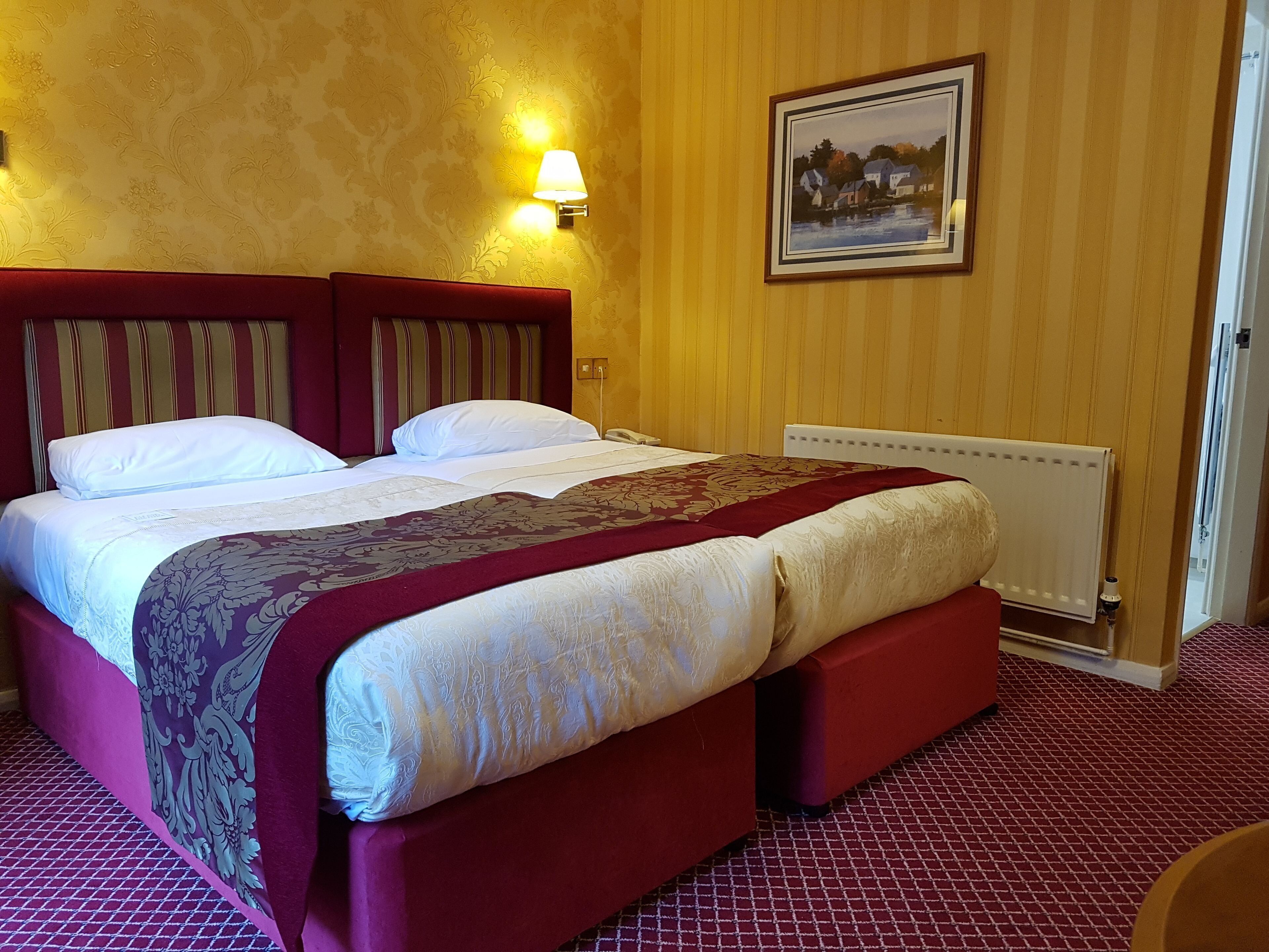 A hotel room with a large bed, a red headboard, a wall-mounted lamp, and a framed picture on the wall.